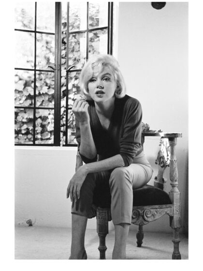 Marilyn Monroe sitzt nach vorn geneigt auf einem ornamentierten Sessel vor einem geöffneten Fenster