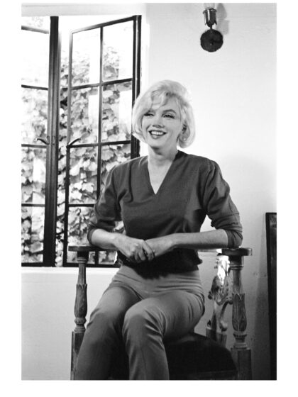 Marilyn Monroe sitzt lachend auf einem Holzstuhl vor einem geöffneten Fenster