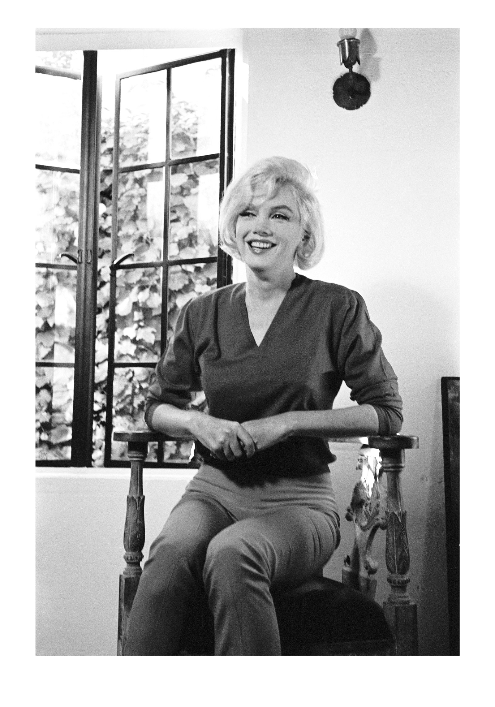Marilyn Monroe sitzt lachend auf einem Holzstuhl vor einem geöffneten Fenster