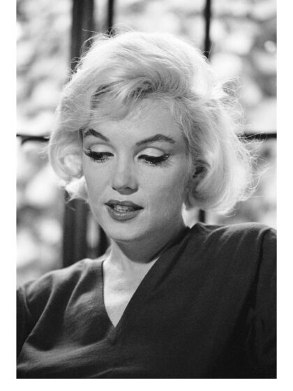 Nahaufnahme von Marilyn Monroe mit leicht geneigtem Kopf und gesenktem Blick vor einem Fenster