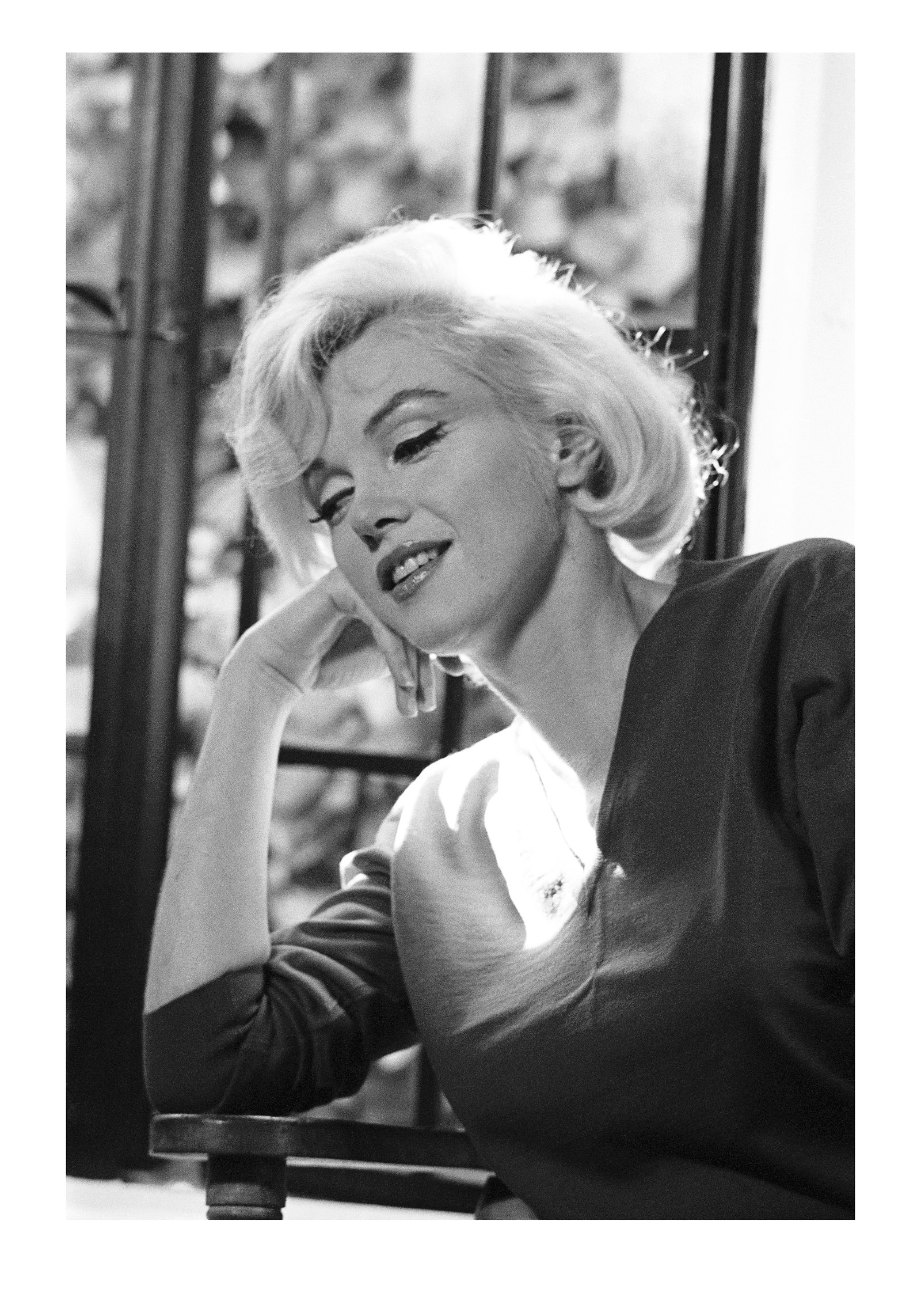 Schwarz-Weiß-Porträt von Marilyn Monroe mit geneigtem, auf die Hand gestütztem Kopf vor einem Fenste